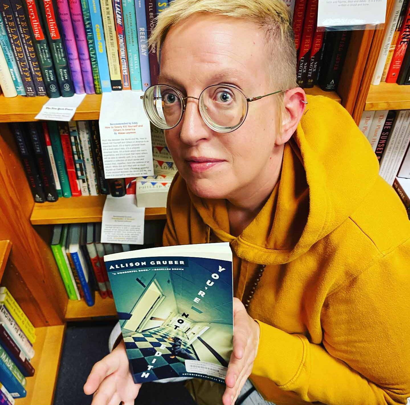 A young woman with thick glasses and a bright yellow hoodie in a bookstore holding the book she wrote