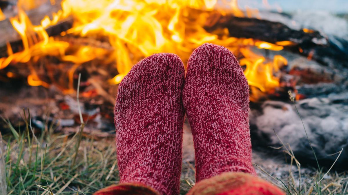 A pair of feet in pink wool socks in front of a campfire during the day.