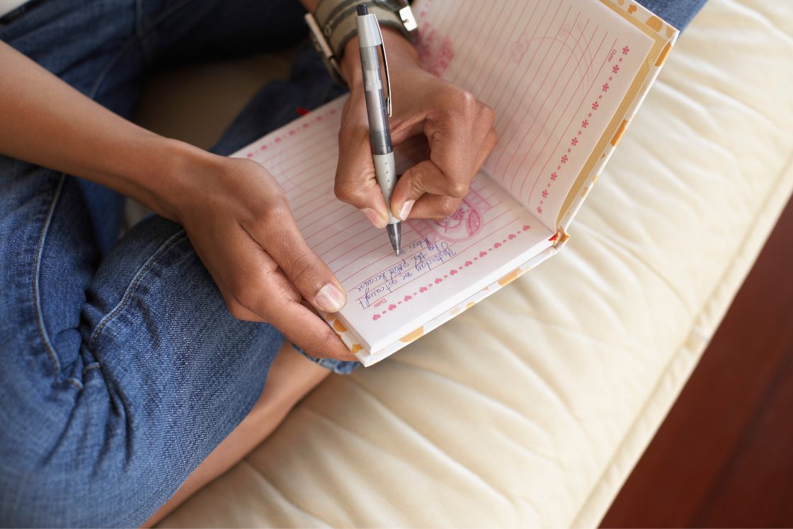 A Black woman's hands write into a journal over crossed legs.