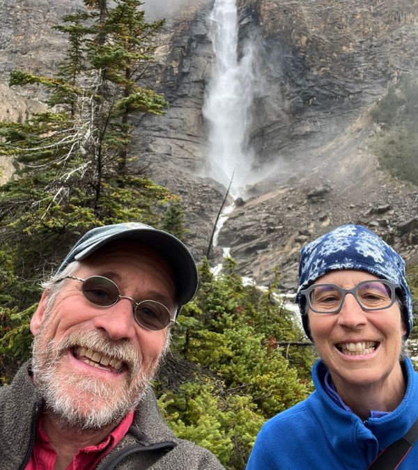 Mary Evelyn Burman and husband take a selfie. A streaming waterfall meanders down a mountain behind them.