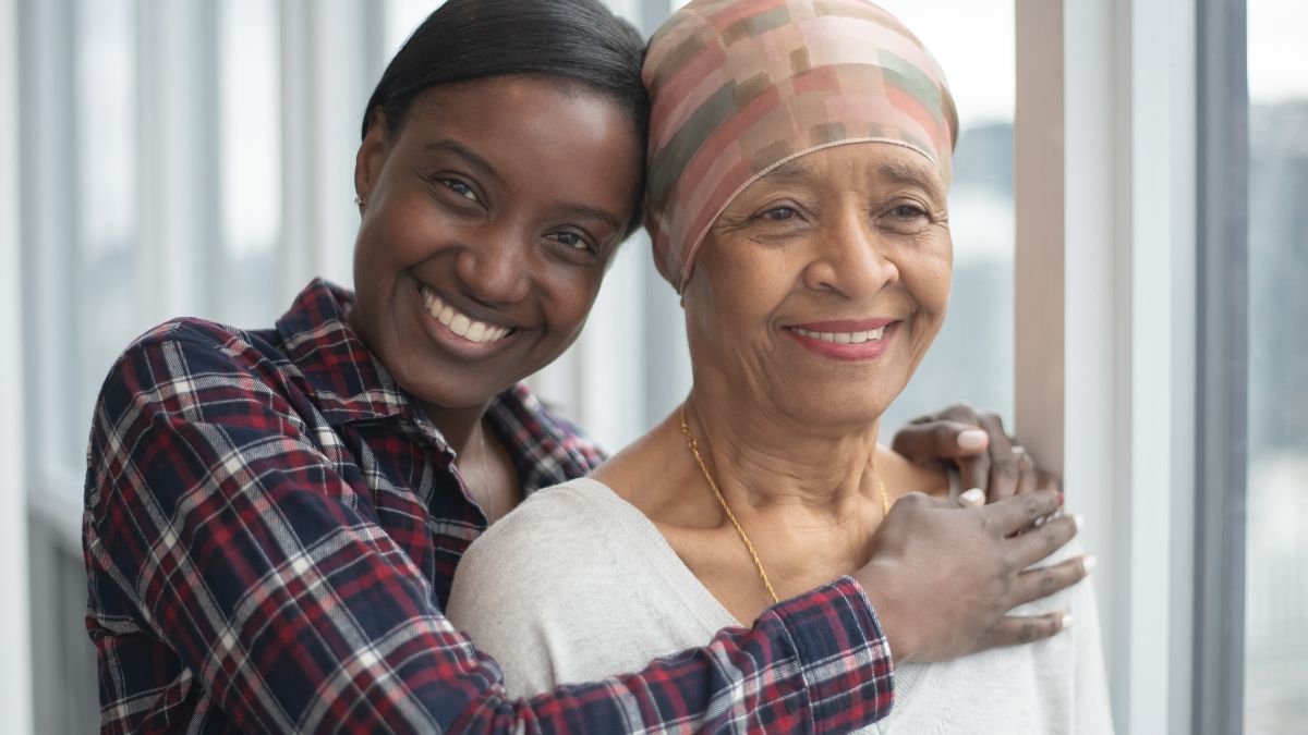 A smiling Black woman wearing a headscarf is embraced by her daughter