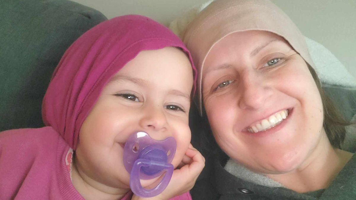 Amy Almas snuggles with her young daughter who has a purple pacifier in her mouth. They both wear soft knit caps on their heads.