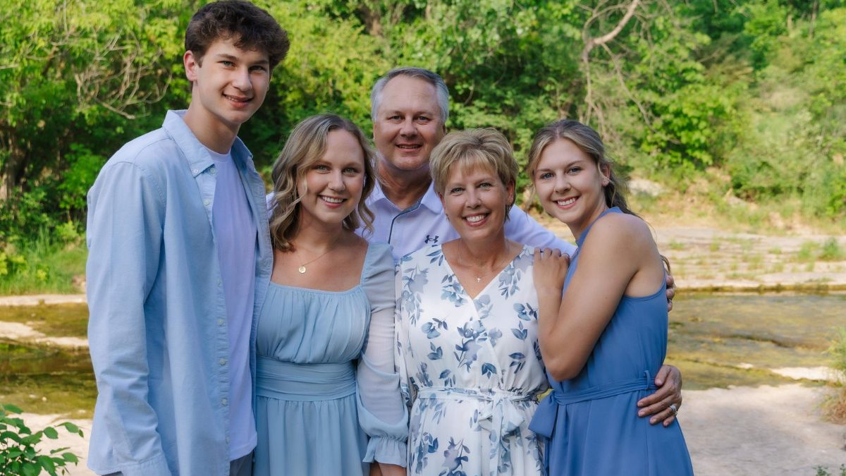 Tammy Uhl is surrounded by her husband and their three grown children, all wearing shades of blue and white in a tranquil outdoor setting