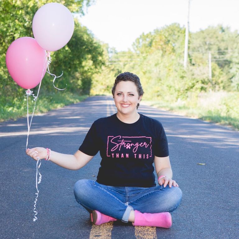 Ashley walls sitting in the middle of the road holding pink balloons. Her shirt reads, "Stronger than this."