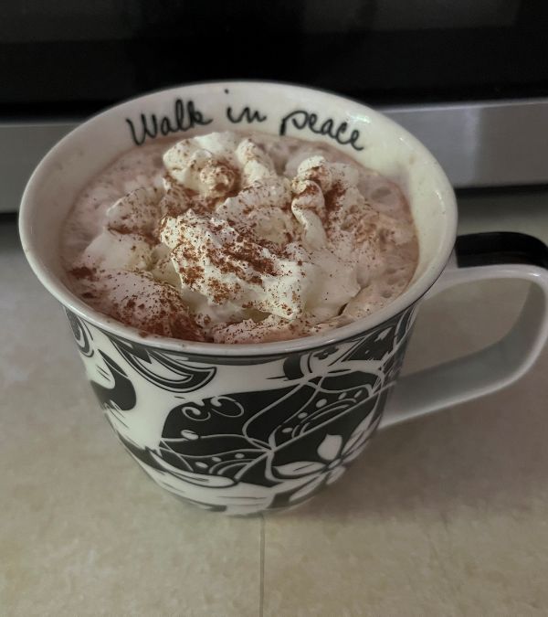 Cinnamon sprinkles the whipped cream atop homemade coffee in a blue and white mug inscribed with "Walk in Peace"
