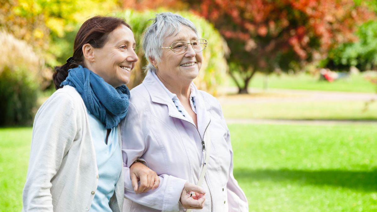 A middle-aged woman walks with her elderly mother amidst a beautiful fall landscape.