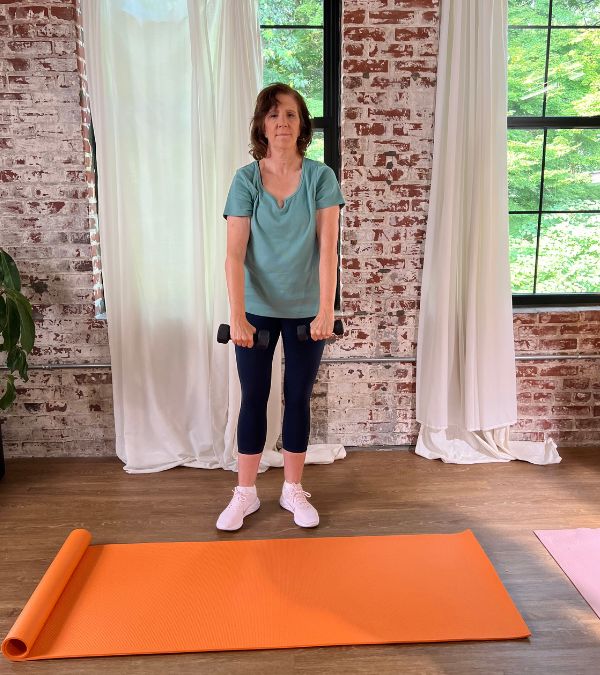 Monica Steigerwald stands before an orange yoga mat, lifting weights.