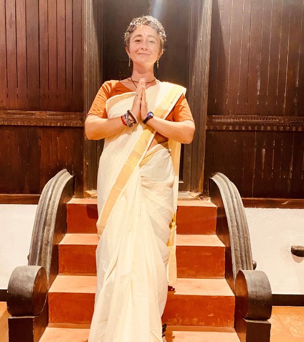 Wearing a sari, Jenna Antonelli clasps her hands in grateful prayer in front of a dark wood temple.