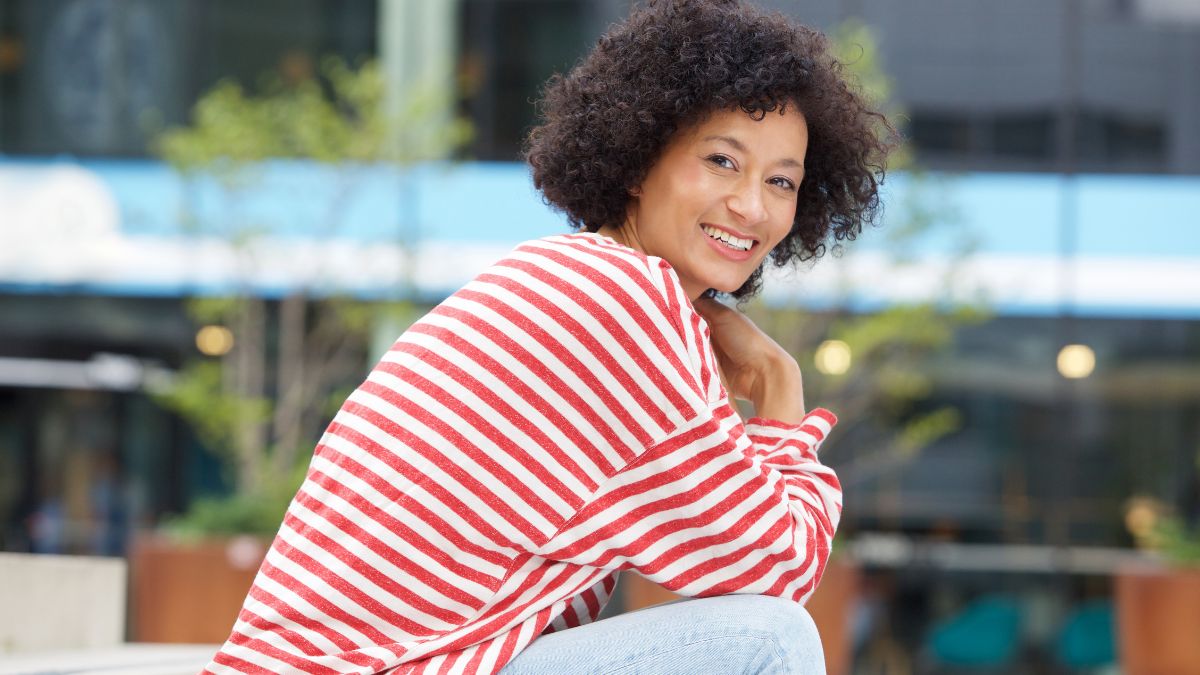 A Black woman turns to display a vibrant smile in her red-and_white sailor top as she sits in an urban setting.