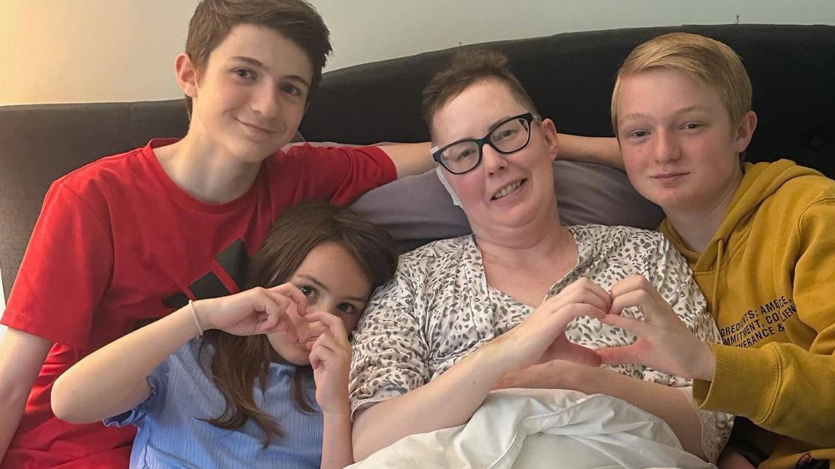 Julie Rock rests and recovers after breast cancer treatment with three of her young children gathered around her making the heart symbol with their hands.