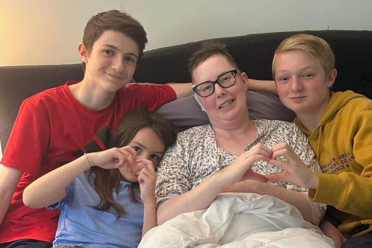 Julie Rock rests and recovers after breast cancer treatment with three of her young children gathered around her making the heart symbol with their hands.