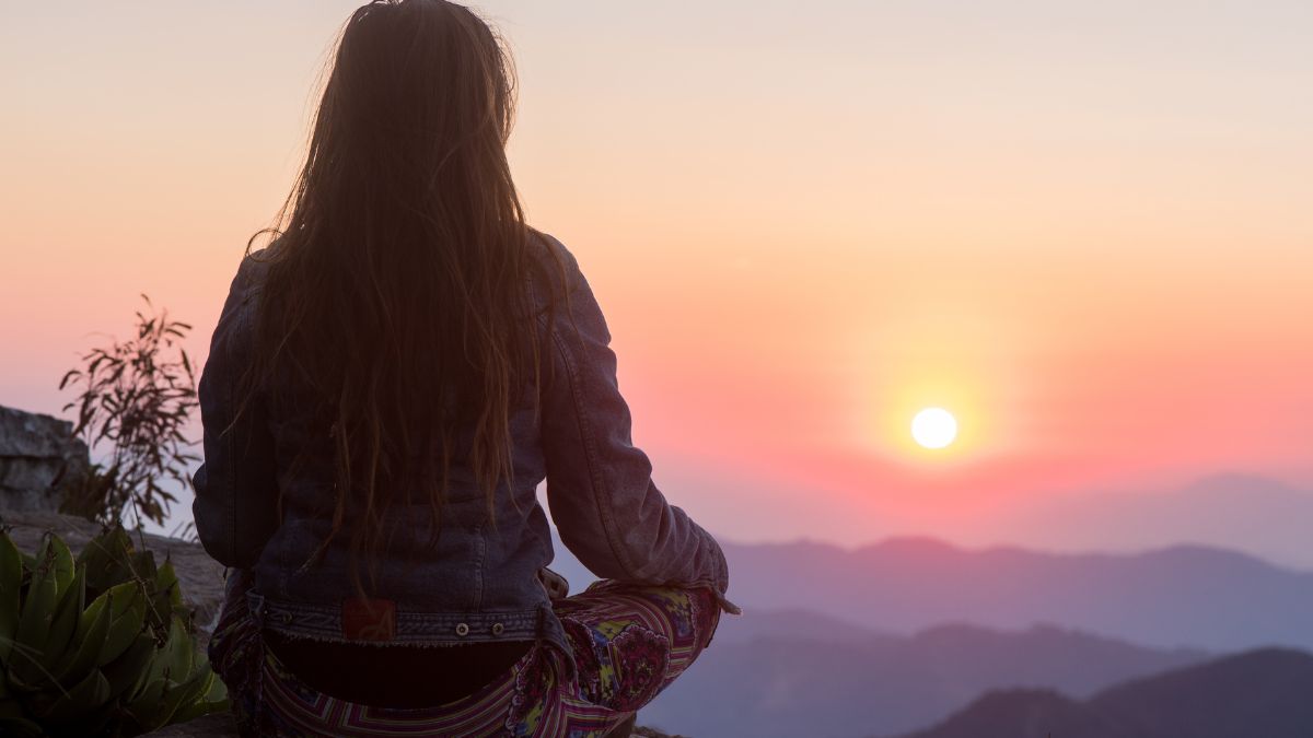 A woman sits with her back towards us watching the sunrise over a colorful ridge of mountains.