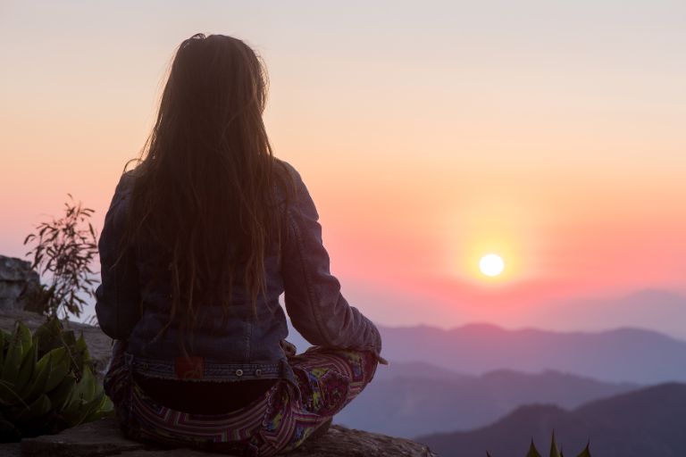 A woman sits with her back towards us watching the sunrise over a colorful ridge of mountains.