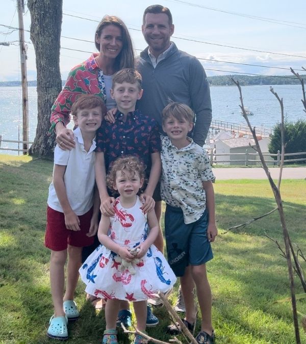 Karen Powell, her husband, and four young children pose in front of beautiful, sunny lake setting.