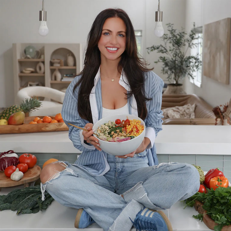 Lainie Kates sitting criss crossed on the kitchen counter, smiling, holding a bowl of veggies.
