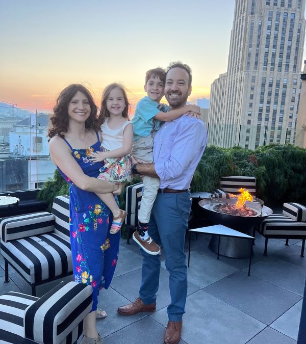 Lilly Ribner holds her daughter as she stands next to her husband, who holds their son, standing on a beautiful hotel rooftop at sunset.