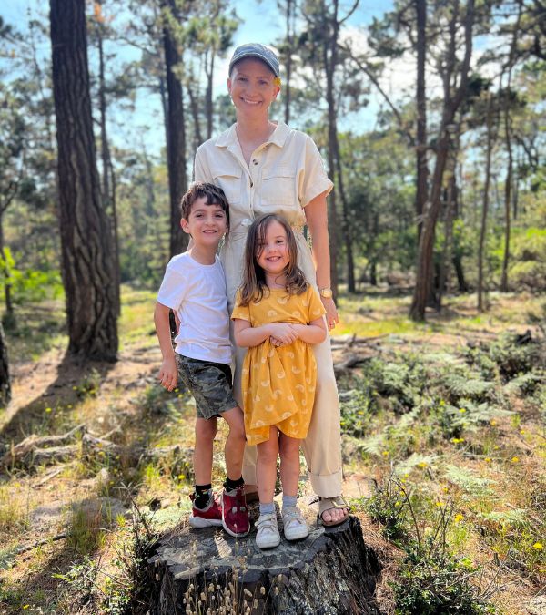 Lilly Ribner stands with her two young children in a beautiful patch of nature, many trees behind them.