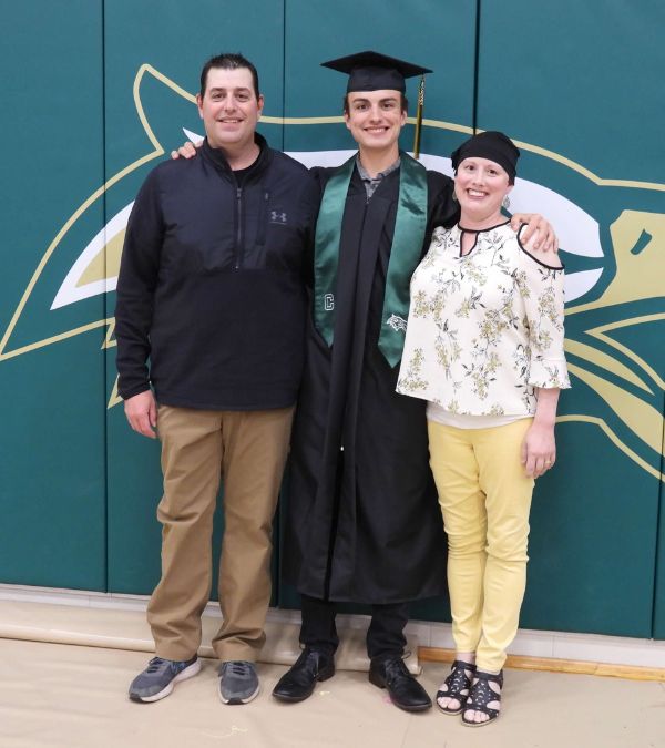 Klara Hartfiel stands proudly with her son, who wears his graduation cap and gown. Her husband stands on the other side of their son.