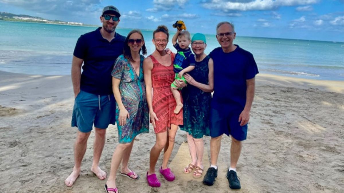 Maura Roby stands with her husband and two daughters' families on the beach, tranquil blue ocean behind them.