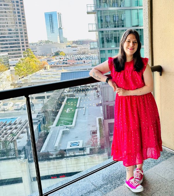 Molly Gaynor stands by a hotel balcony, high above street level, an interesting urban scene below, but the main interest is Molly's pink dress and radiant smile.