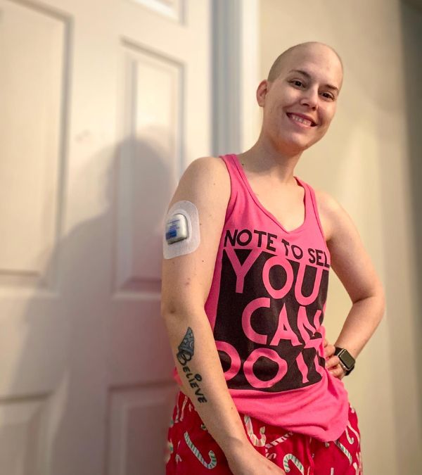 Molly Gaynor stands with her head shaven, wearing bright pink breast cancer walk/run gear, a happy smile on her face.