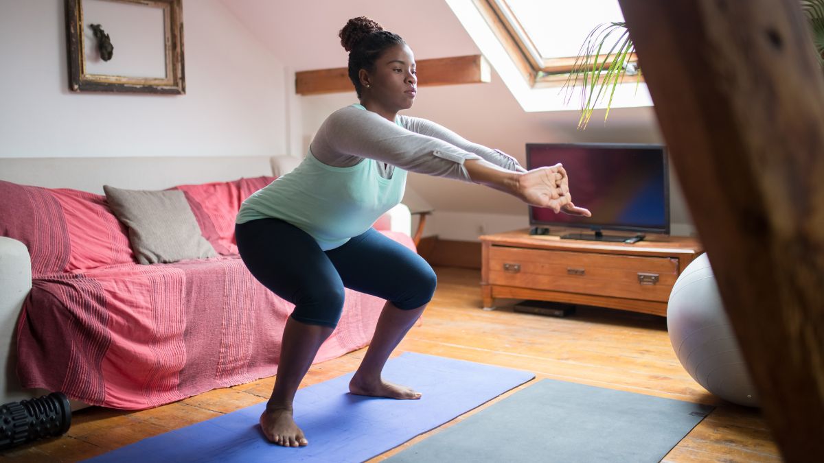 A Black woman exercises in her home by doing a deep squat on her yoga mat.