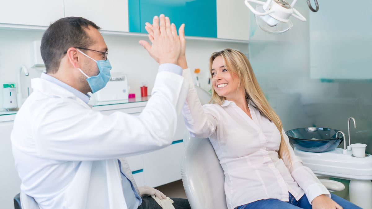 A woman sitting in the dentist's chair slaps a high five with her dentist.