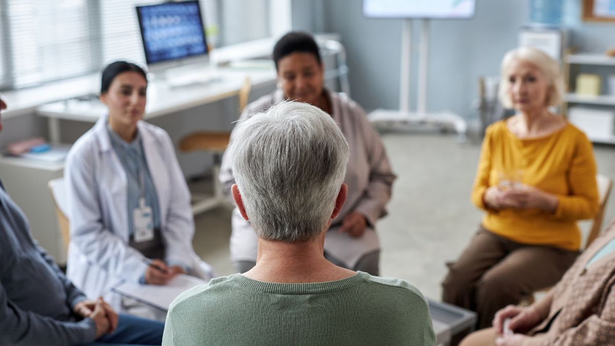 A support group of diverse women gather round a clinical therapist in a comforting circle.