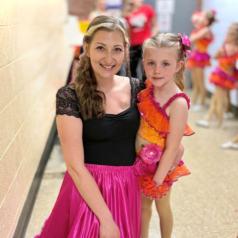 Kara Taylor holds her daughter as they wait in a school hallway for a performance, dressed in vibrant, colorful, and flouncy costumes, suitable for a dance.