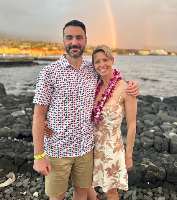 A rainbow shimmers behind Lisa Csensits and her husband as they stand on an island beach.