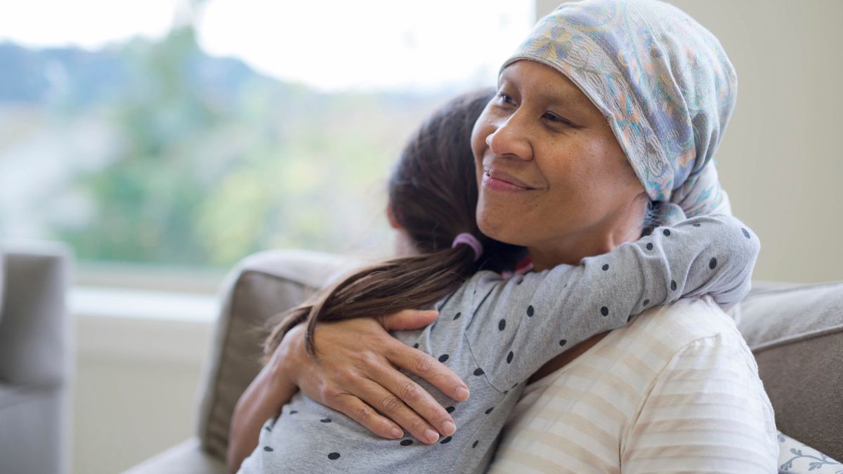Seated in an armchair, a woman, who is wearing a headscarf and impacted by cancer, is hugged meaningfully by her granddaughter.