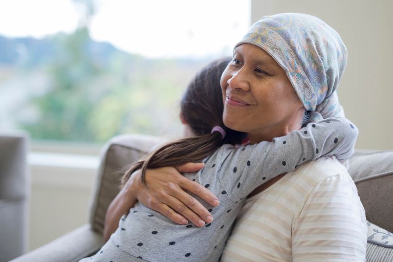 Seated in an armchair, a woman, who is wearing a headscarf and impacted by cancer, is hugged meaningfully by her granddaughter.