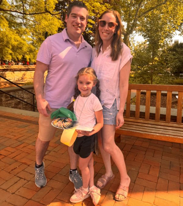 Melissa Berman stands with her husband and daughter in a beautiful outdoor setting during summer.