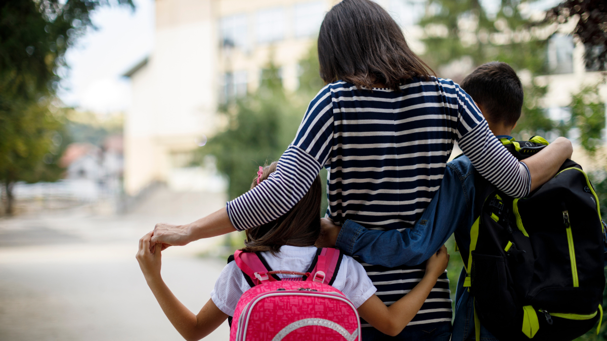 A Black woman with her arms around her son and daughter, walking them into school
