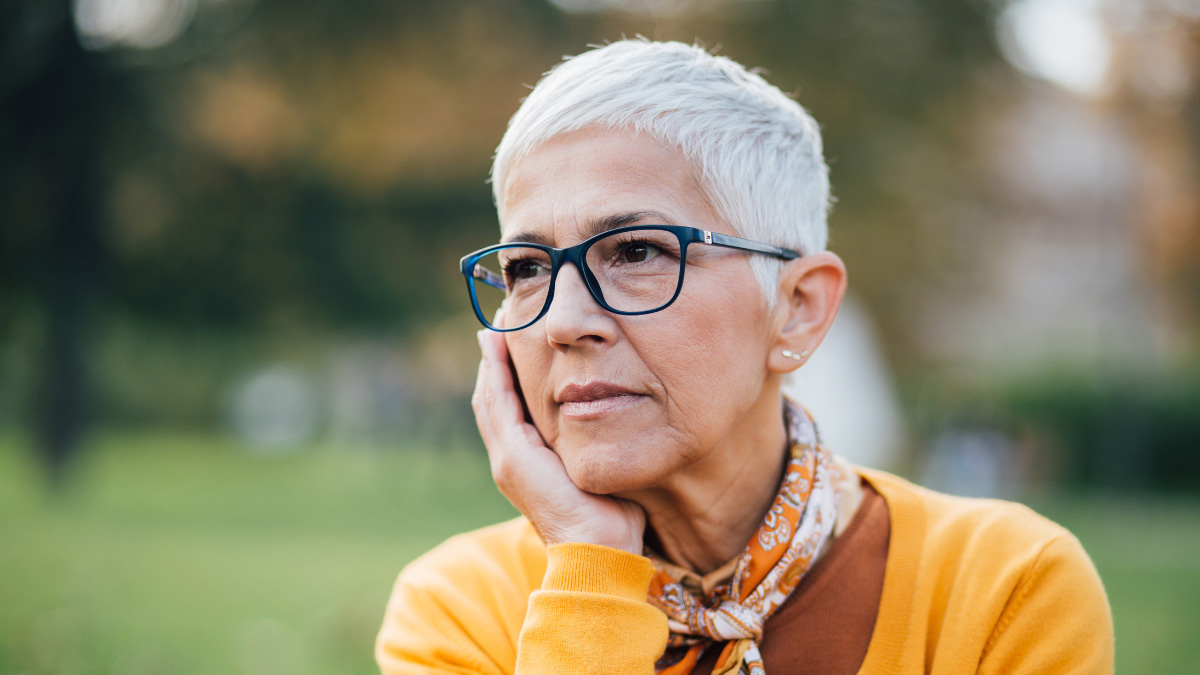An older white woman with glasses looks thoughtfully into the distance, resting her head in her hand.