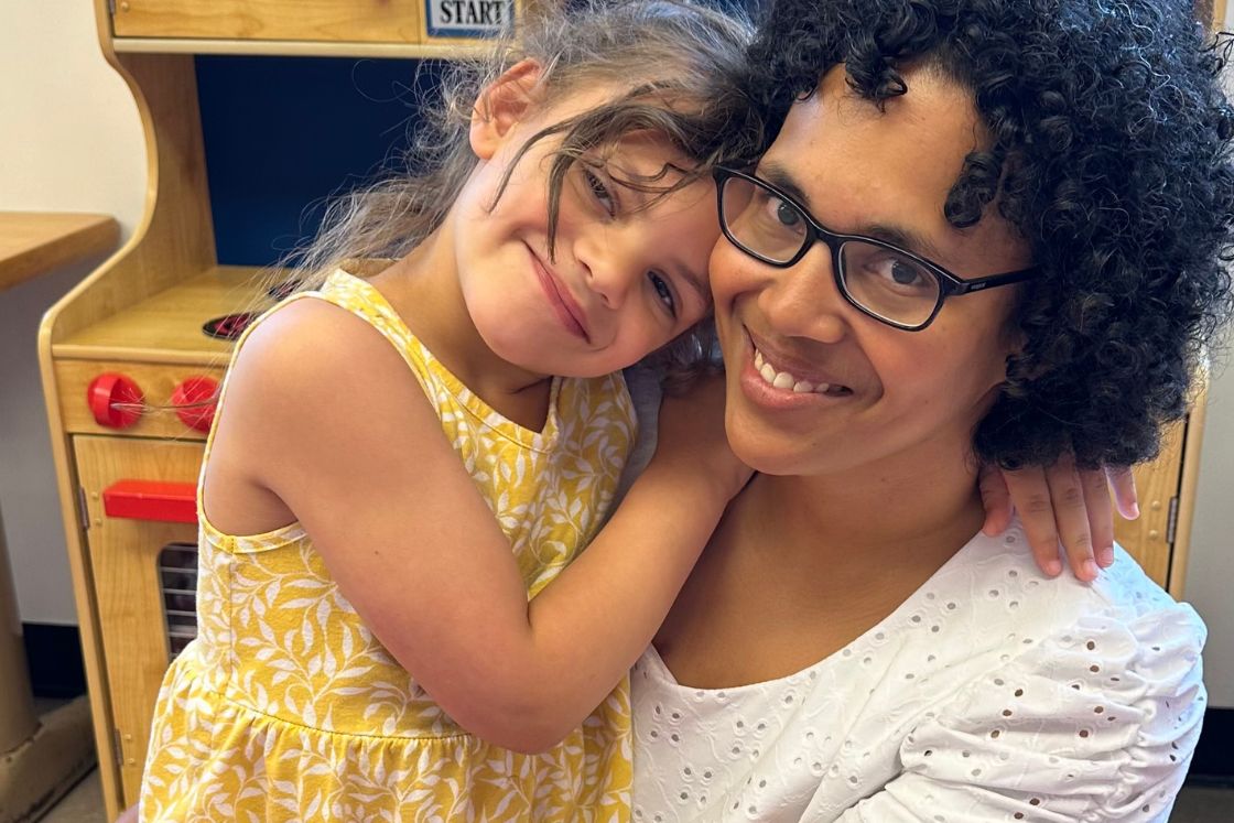 Angel Rinker holds her young daughter who cuddles close to her neck with a big smile.