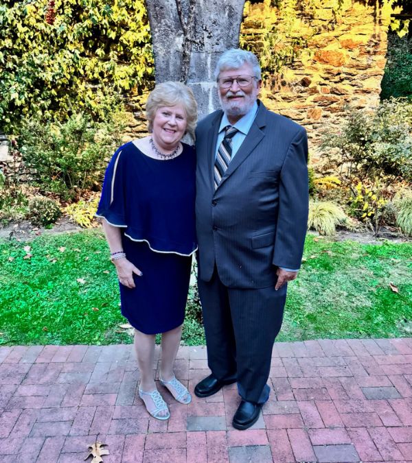 Jeanmarie Mason and her husband Don are elegantly dressed for a formal event as they stand in front of a yellow forsythia bush.