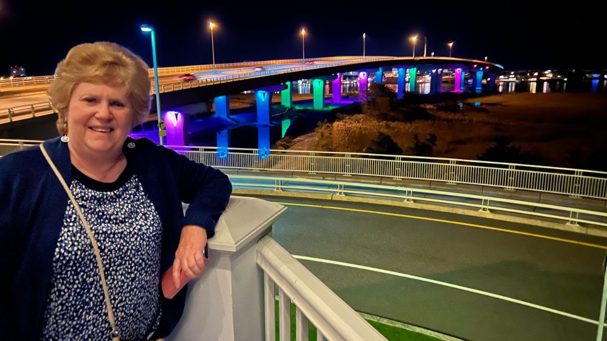 Jeanmarie Mason stands before the Ocean City bridge which is lit up at night in the MBC Awareness colors.