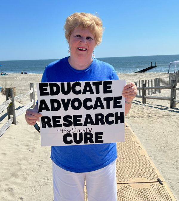 Jeanmarie Mason stands with an advocacy sight for MBC: Educate, advocate, research, cure, as her 4 words for Stage IV breast cancer.