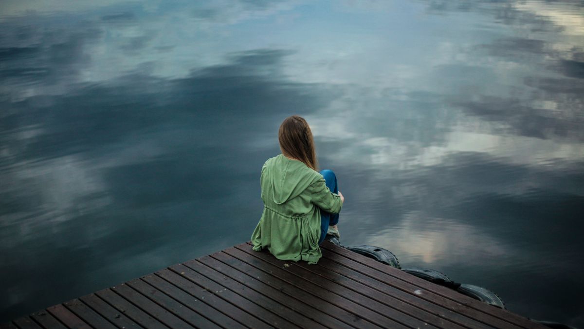 A young woman sits on a dock looking thoughtfully at the lake ahead of her.