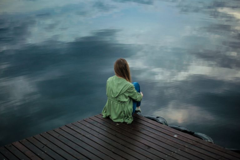 A young woman sits on a dock looking thoughtfully at the lake ahead of her.