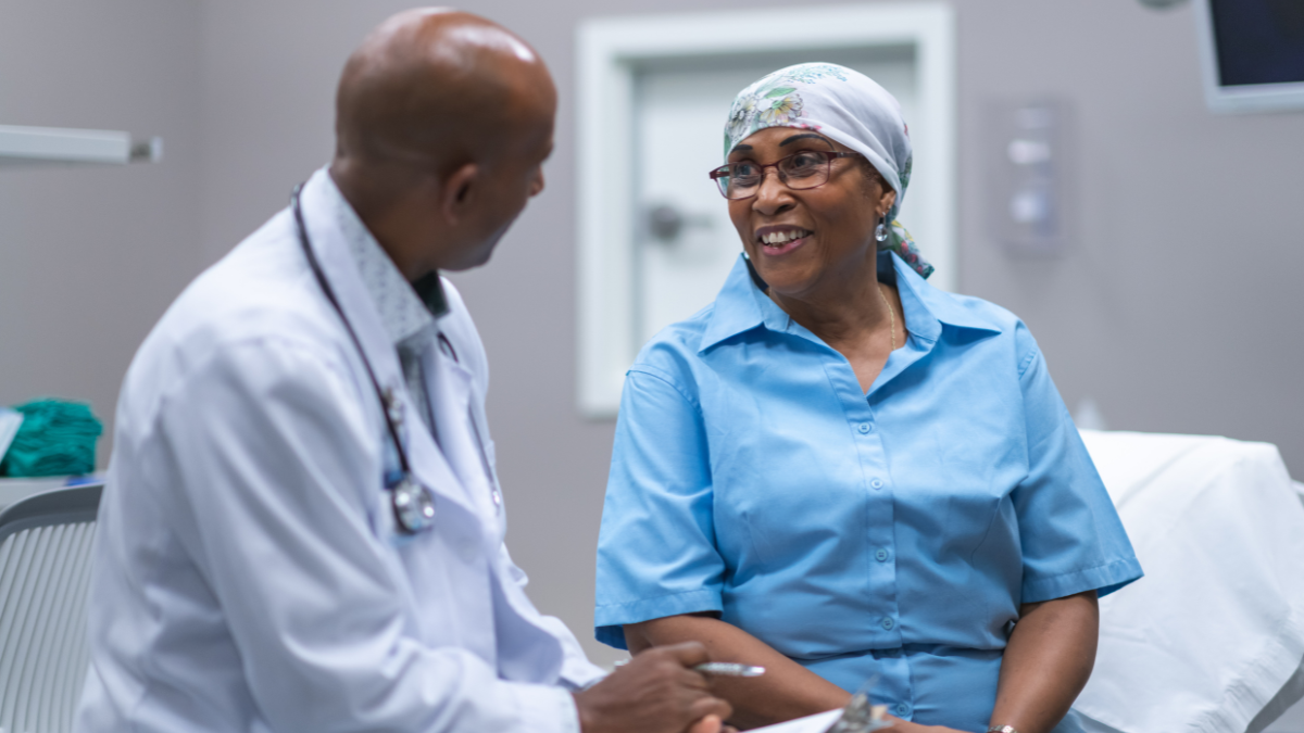 A Black woman sitting on the table at the doctor's office, in conversation with her Black male doctor