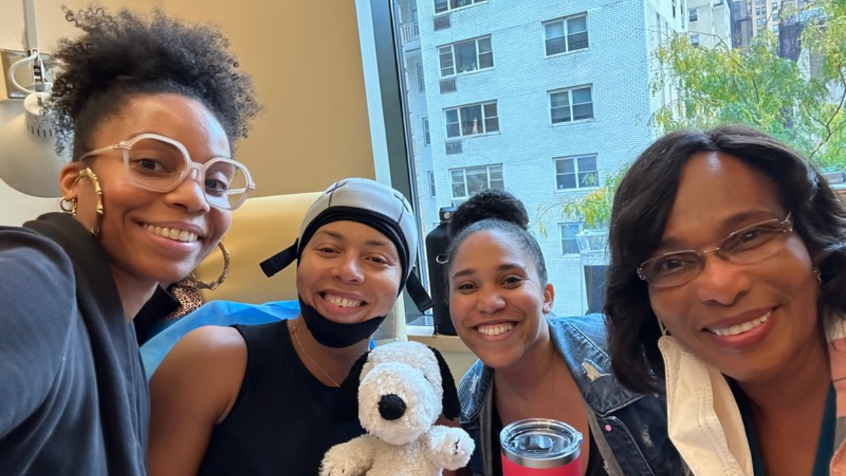 Kia Lee smiling in a chemo chair with a scalp cooling cap on and three family members are smiling around her.