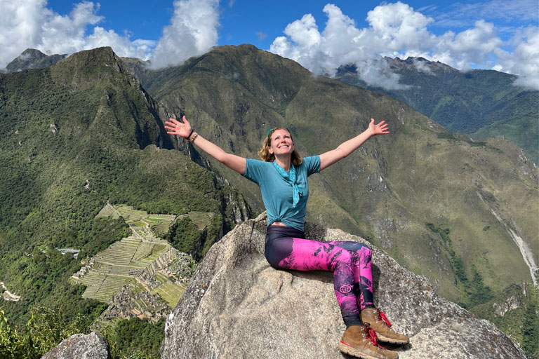 Claudia De Pasquale sitting on top of a rock with her arms out, smiling, on top of Machu Picchu.