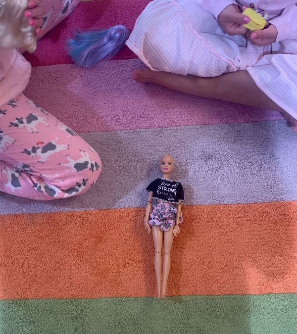 A bald barbie doll lays on a striped rug surrounded by two little girls