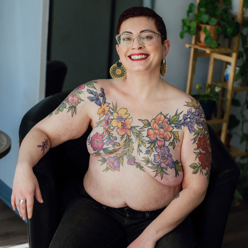 Dani Bennov seated and smiling shows her post-mastectomy floral tattoo covering her chest, shoulders and upper arms.