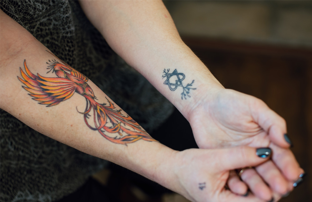 Rebecca shows her forearms, one with a phoenix tattoo and another with a stylized heart tattoo representing her children.