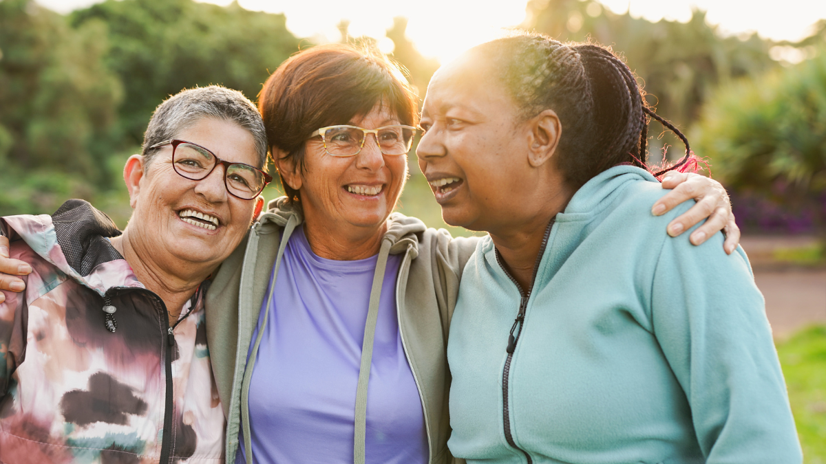 A group of three older women smiling; the one in the center has her arms stretched around the other two