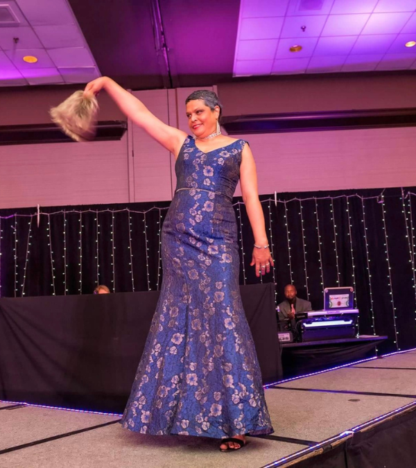 Betty Sanchez removing her wig on stage at a fashion show while modeling a blue evening gown.
