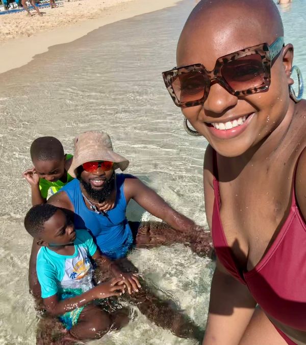Jonise Louis takes a selfie as her family plays in the the crystal clear water of a tropical setting behind her.